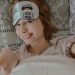 Top view of smiling female reaching out hands to camera while lying on bed under blanket and looking at camera