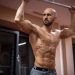 A bald, muscular man performing exercise on gym equipment indoors.